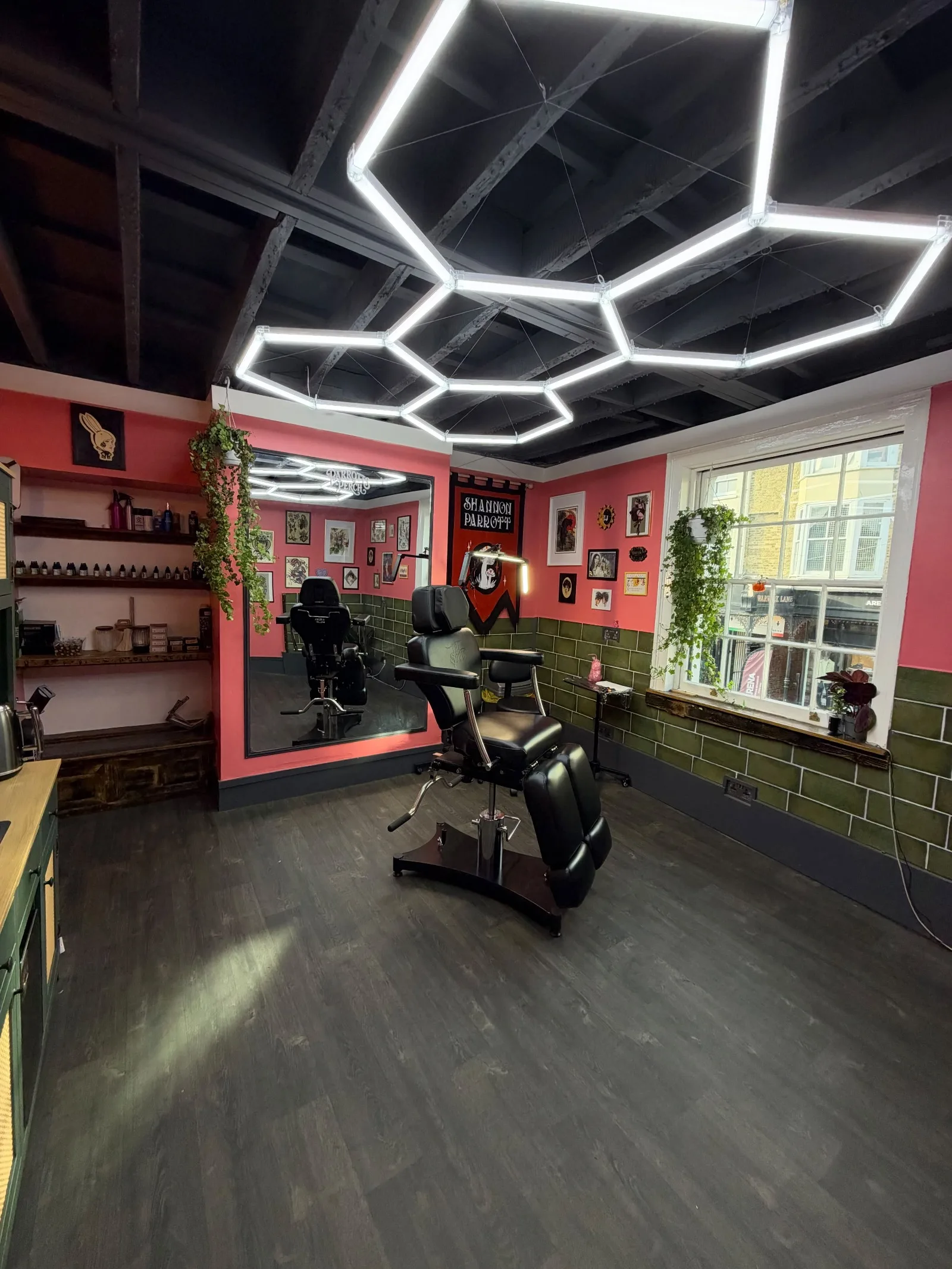 Wide view of the studio: the tattoo chair under hexagon lights, a tall mirror, shelves of ink, the Shannon Parrott banner and a window onto the street