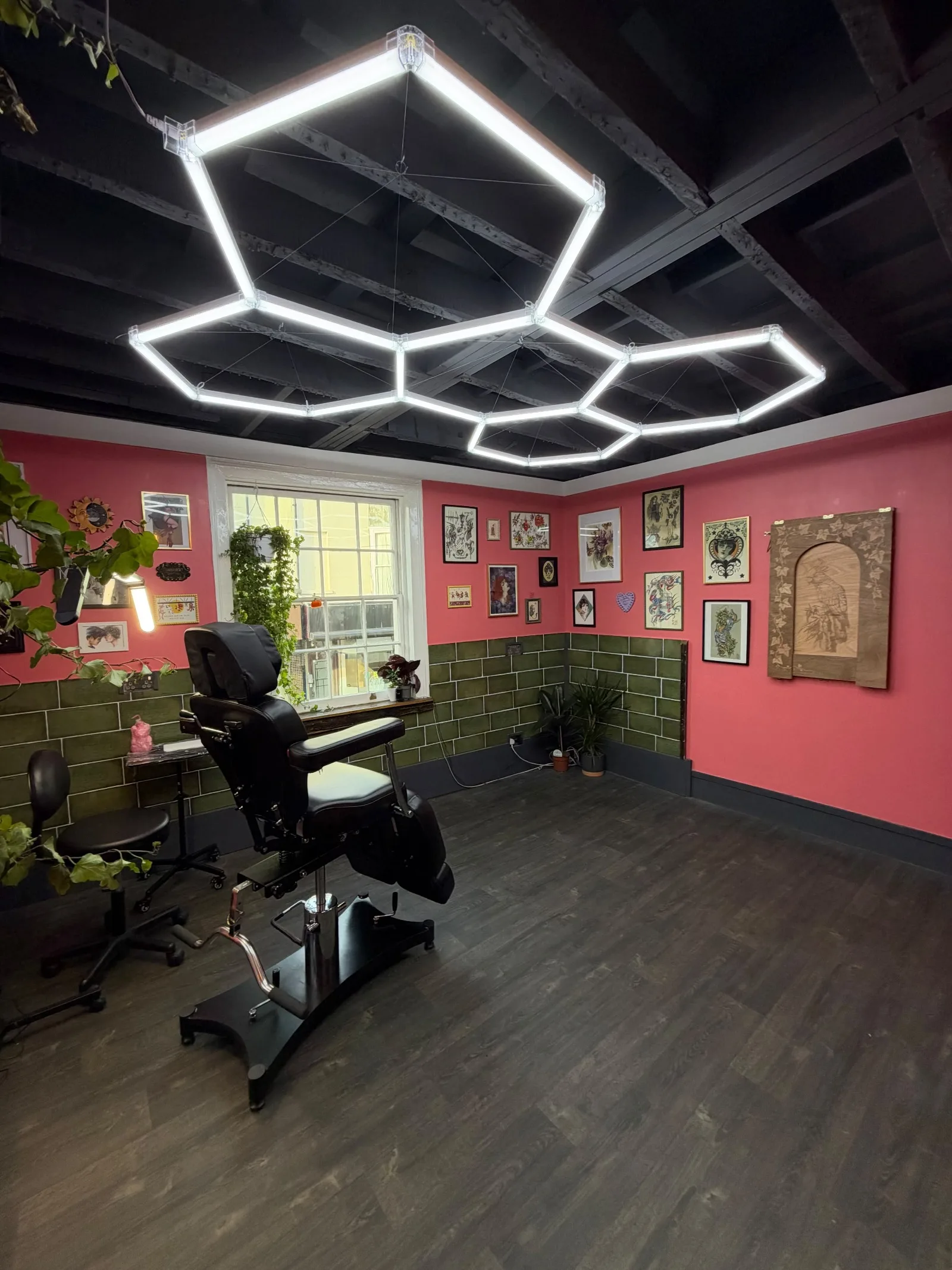 The studio with the black tattoo chair facing the window, framed flash on the pink walls above green tiles, and an engraved wooden cockatoo panel