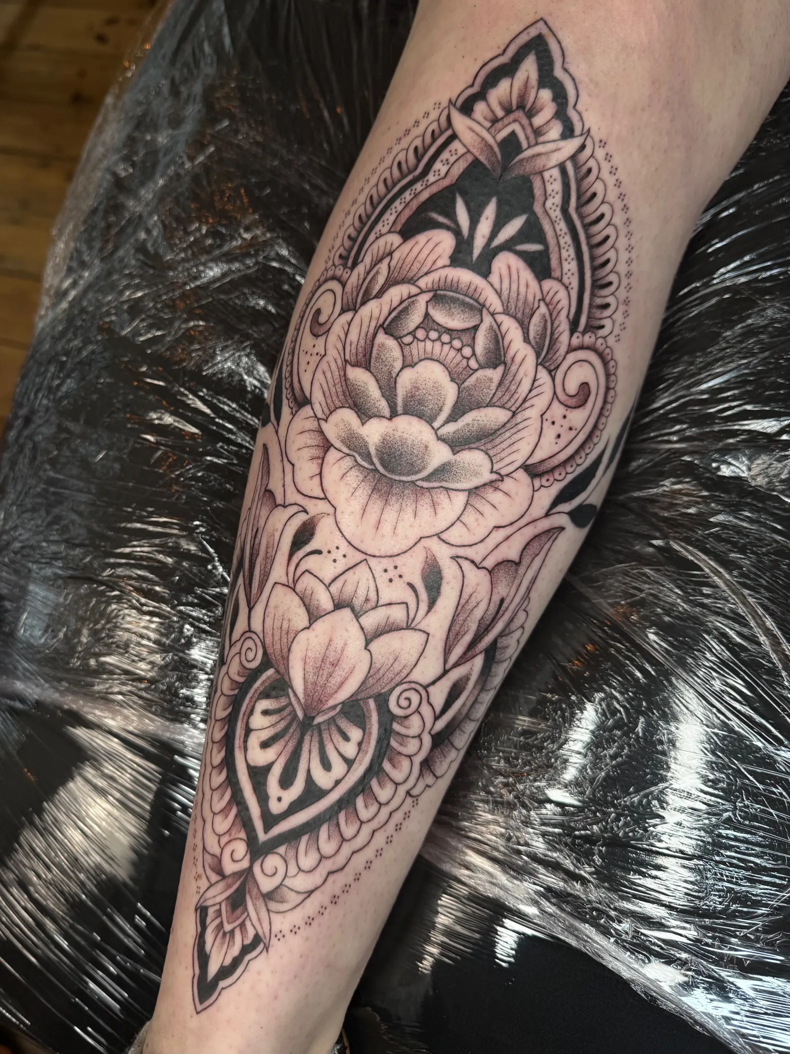Ornamental tattoo on the shin: shaded peonies framed by bold blackwork and fine dotwork patterns