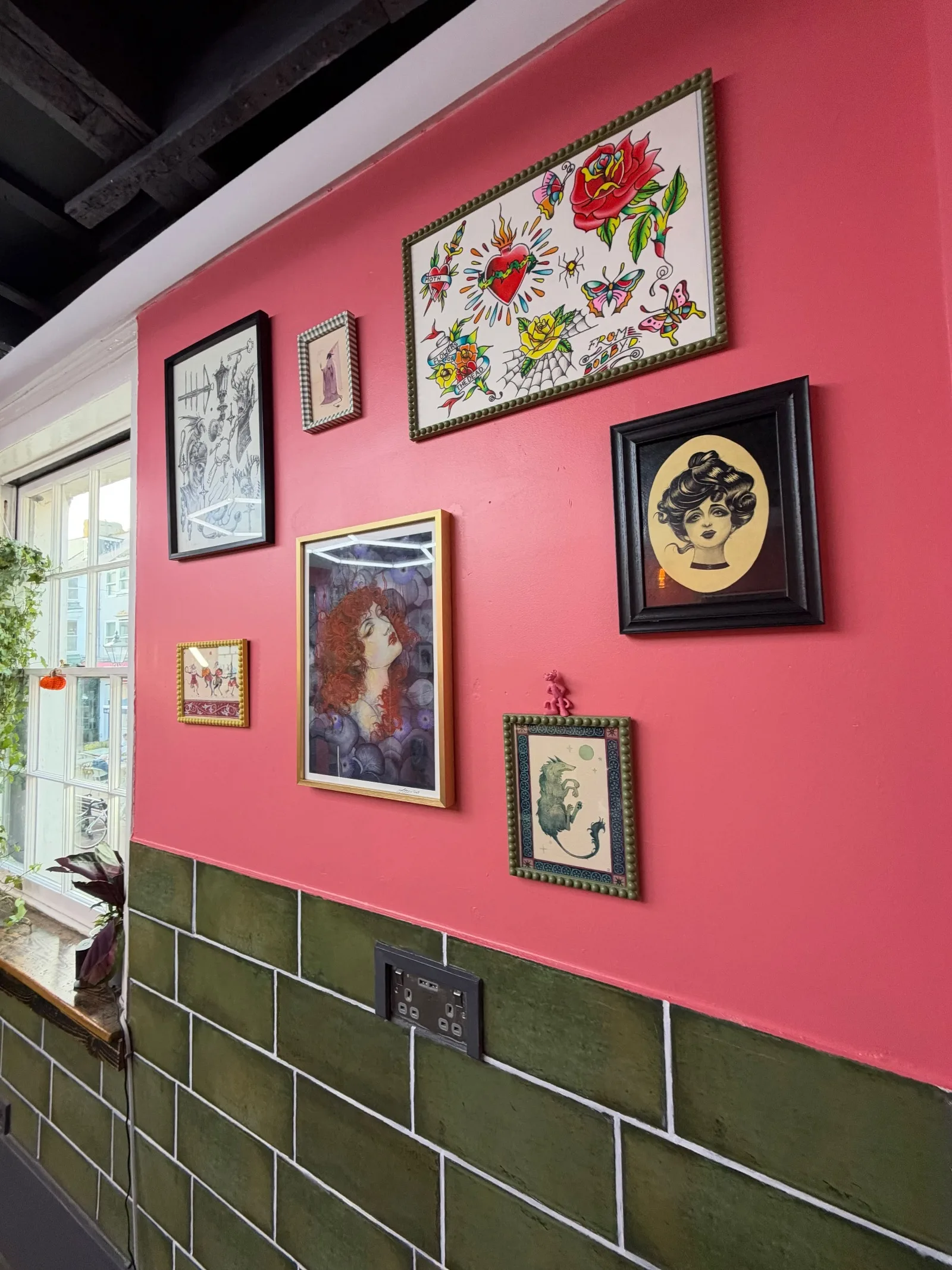 Framed art on the pink studio wall above green tiles: a colourful flash sheet of roses, hearts and butterflies, a red-haired woman, and a Victorian lady in an oval frame