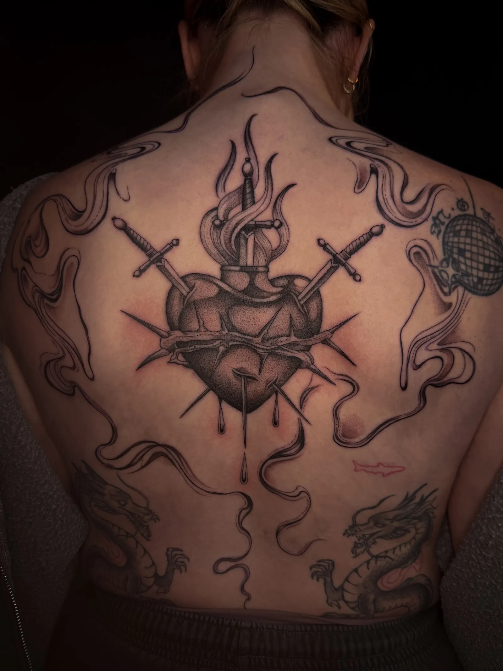 Black and grey sacred heart tattoo across the upper back: a flaming heart pierced by three daggers and wrapped in thorns, with smoke curling out to the shoulders