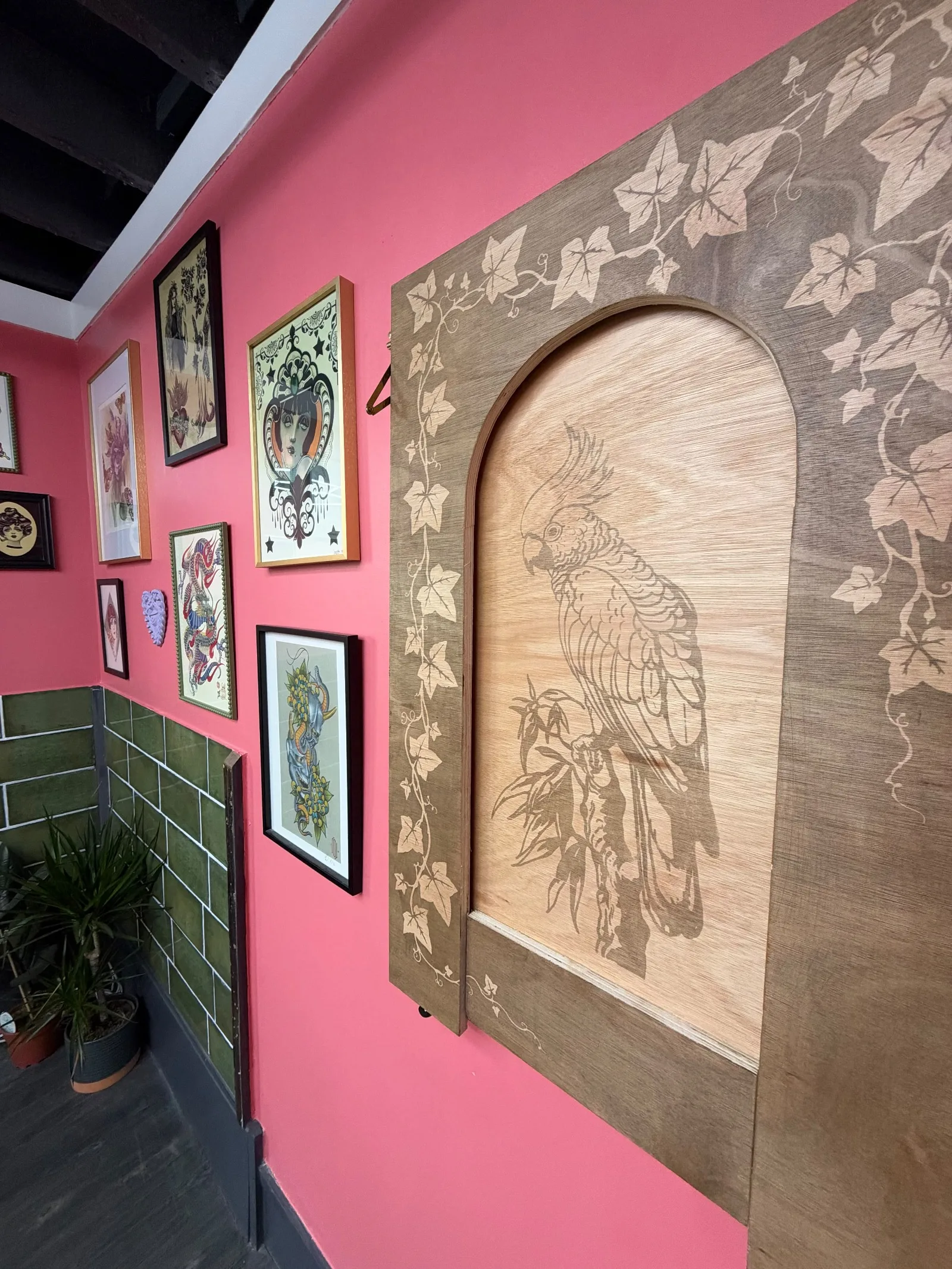 An engraved wooden panel of a cockatoo on a branch, framed by ivy, beside framed tattoo flash on the pink studio wall